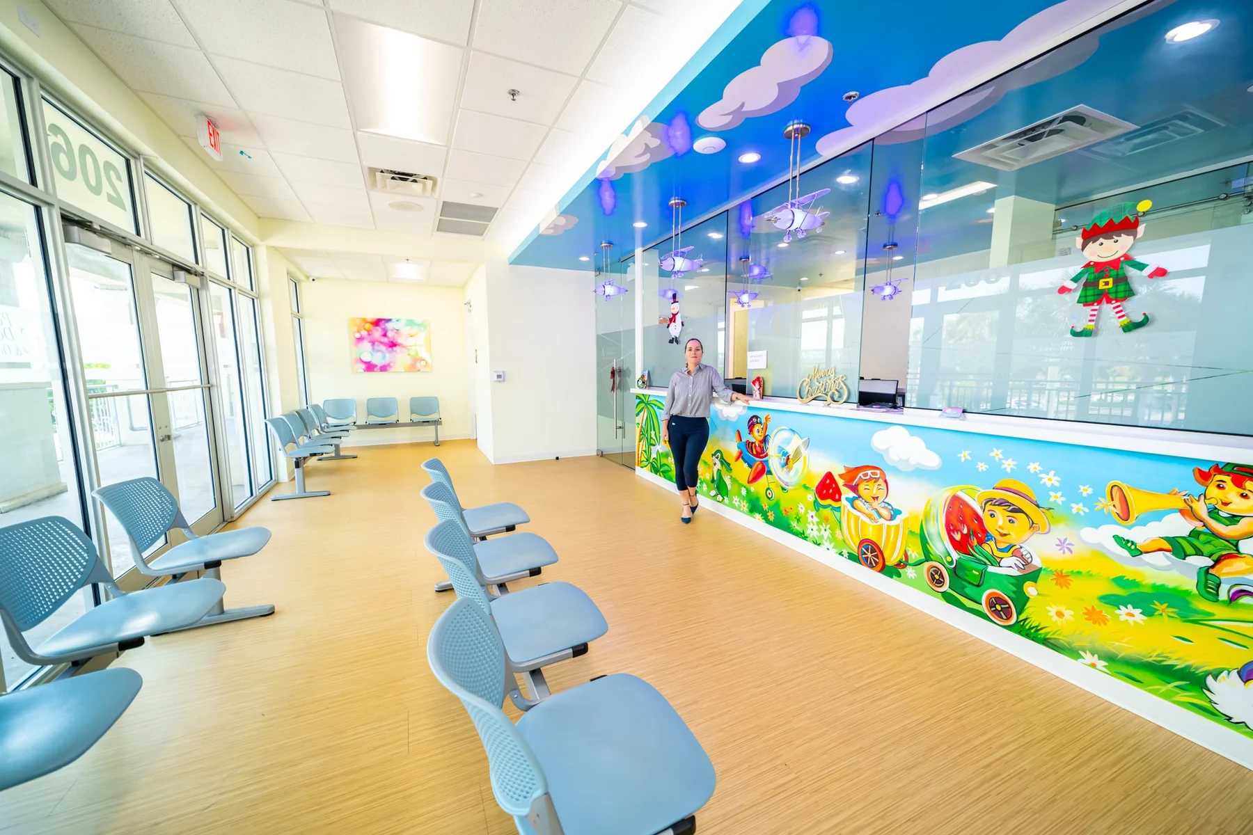 AC Pediatric Dentistry & Orthodontics — Wide view of the waiting room with a person at the mural-painted counter under airplane-shaped lamps