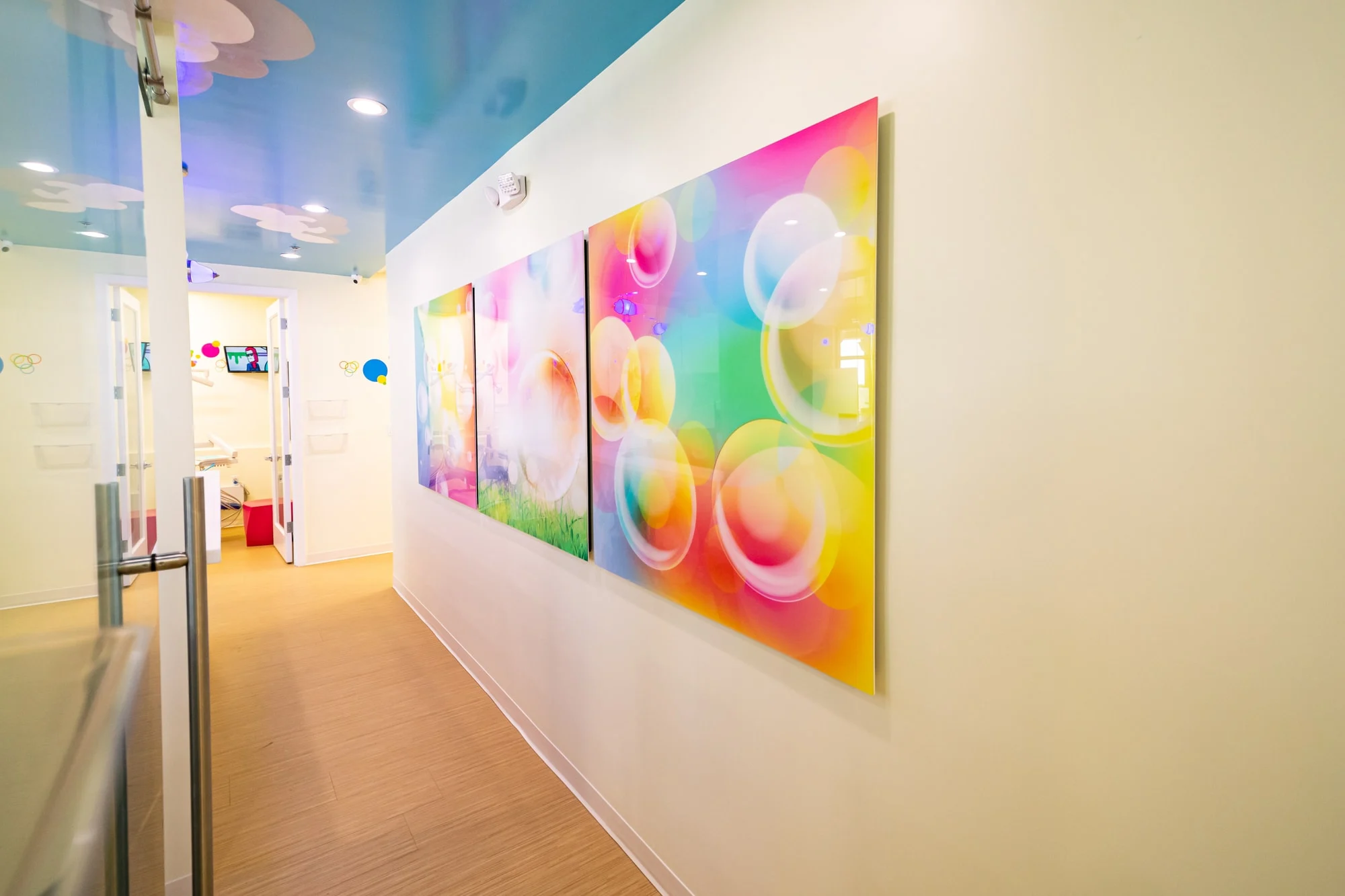 AC Pediatric Dentistry & Orthodontics — Hallway with colorful bubble art panels and a blue ceiling, leading to a treatment room with its doors open