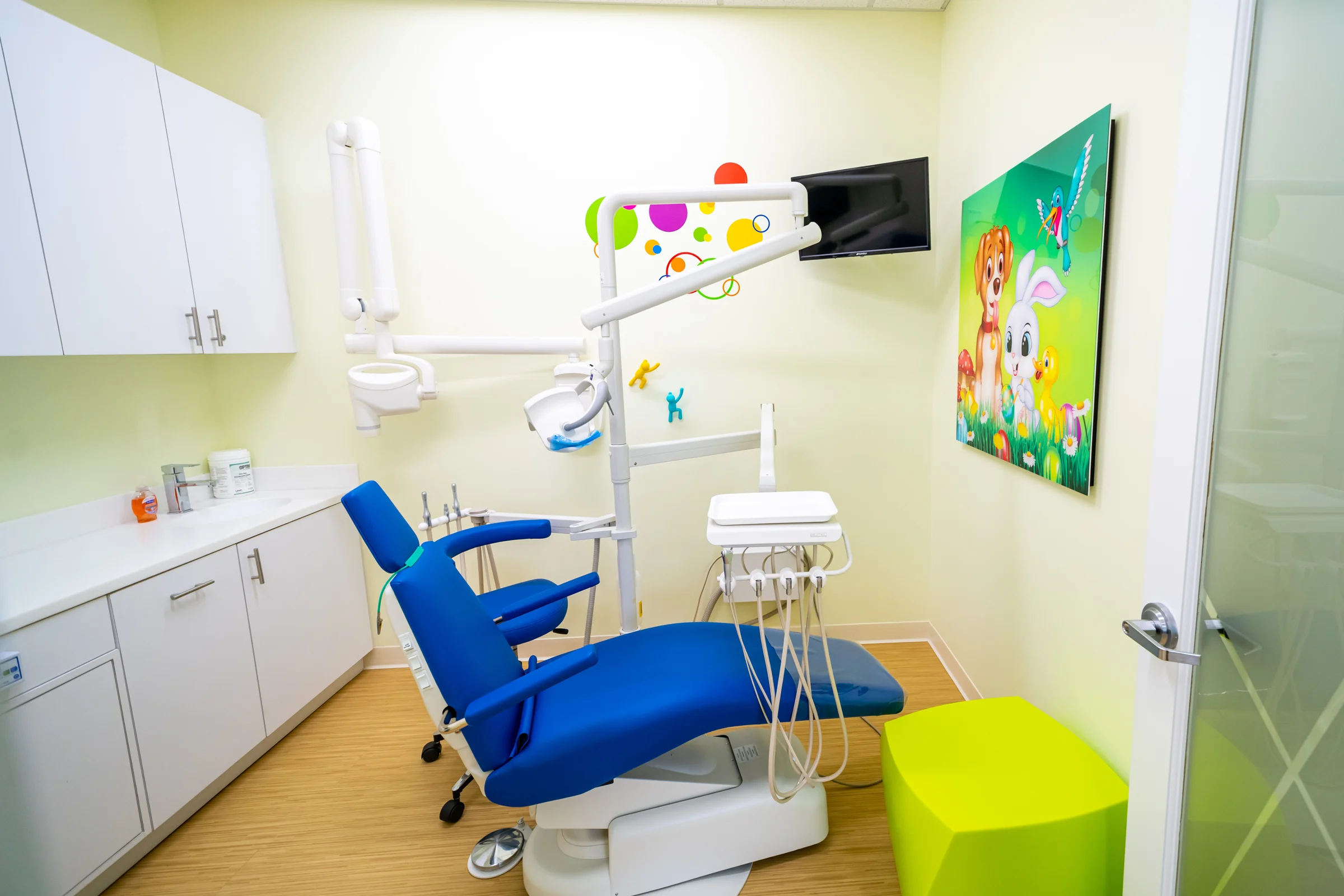 AC Pediatric Dentistry & Orthodontics — Treatment room with a blue dental chair, a wall TV and an art panel of a puppy, a bunny and a duckling