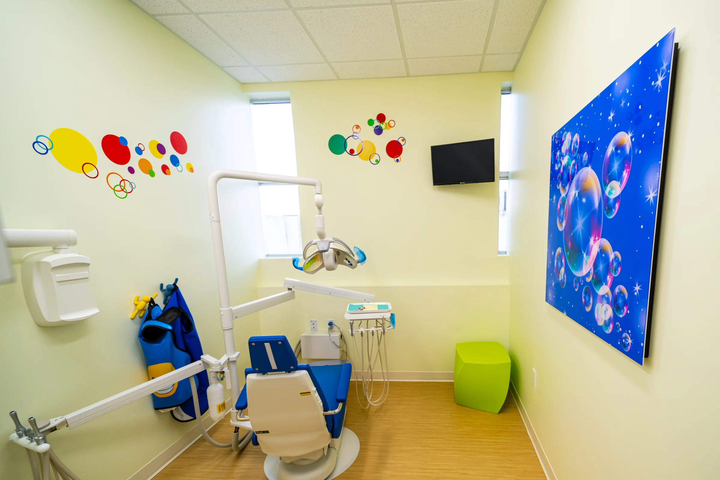 AC Pediatric Dentistry & Orthodontics — Treatment room with a large blue bubble art panel, colorful dot decals, a blue dental chair and a lime-green stool