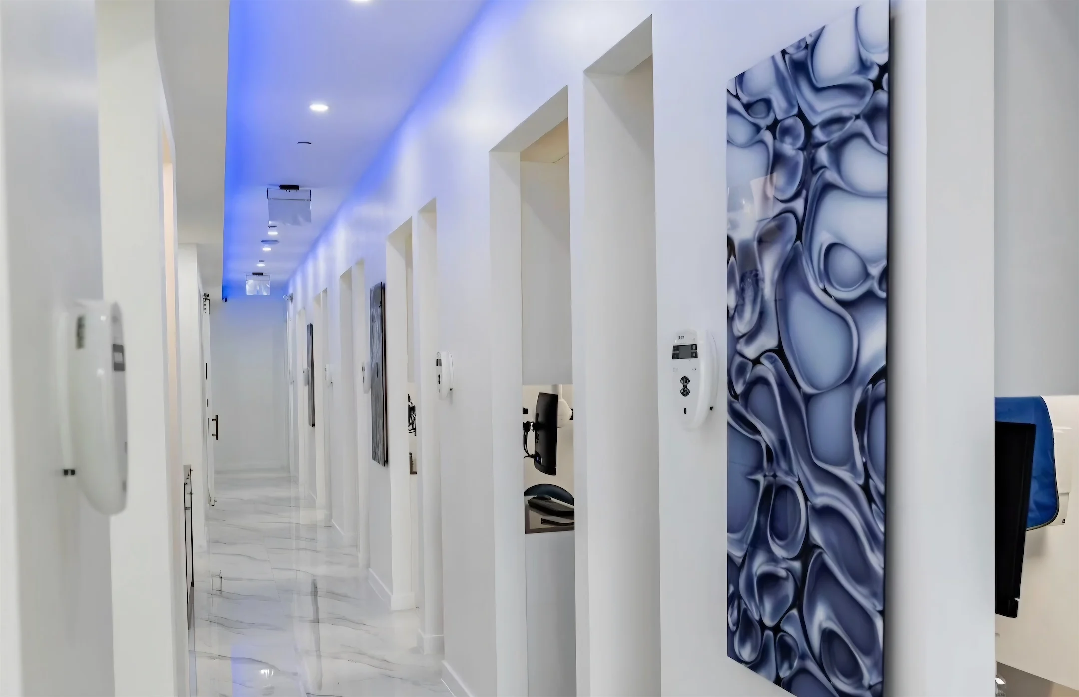 Anillo Dentistry — Pasillo de entradas a las salas de tratamiento bajo una cornisa de luz azul, con un gran panel abstracto azul en primer plano