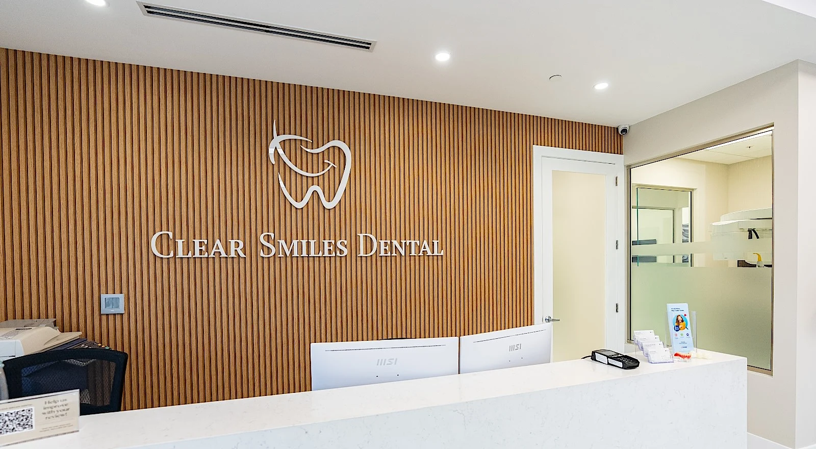 Clear Smiles Dental — Pared de recepción de listones verticales de roble con el logo y el nombre en blanco, detrás de un mostrador de piedra clara