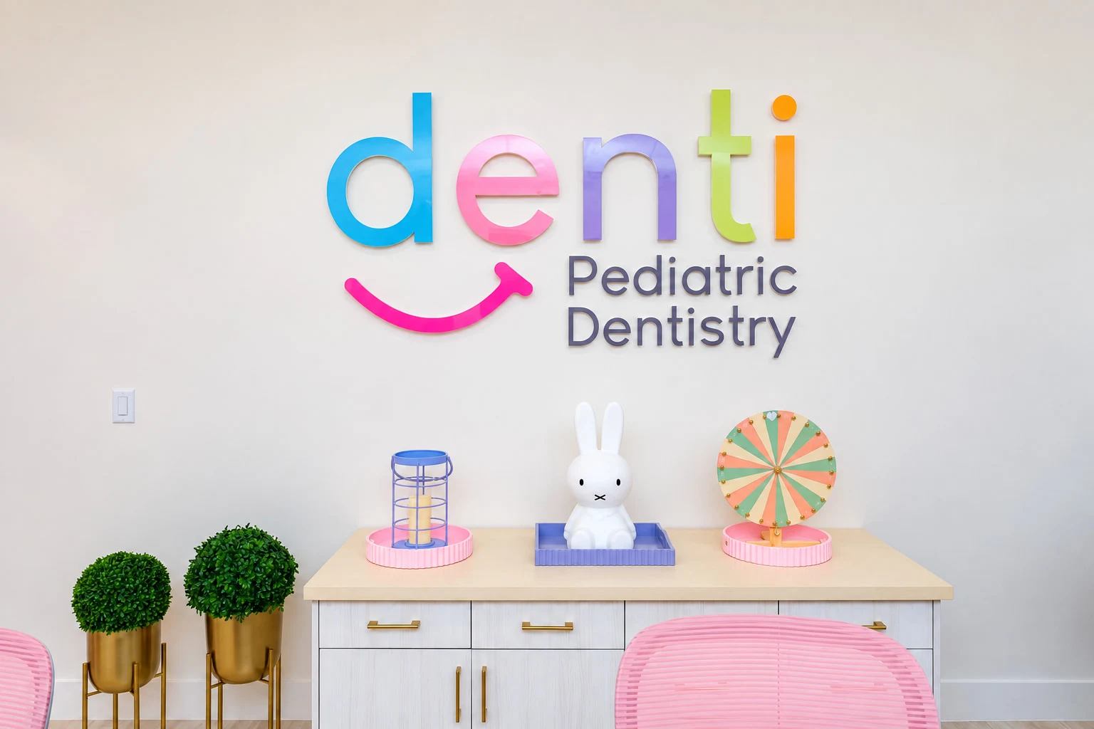 Denti Pediatric Dentistry — Wall with the multicolored “denti Pediatric Dentistry” logo over a cabinet with a white bunny and pink chairs