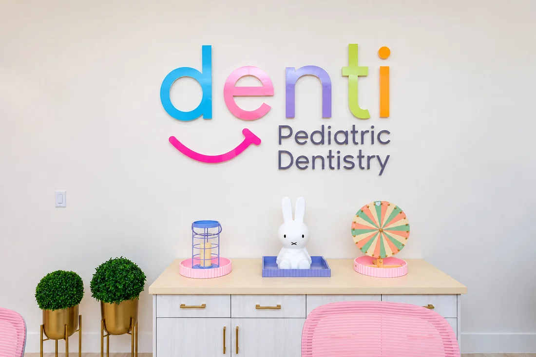 Denti Pediatric Dentistry — Wall with the multicolored “denti Pediatric Dentistry” logo over a cabinet with a white bunny and pink chairs