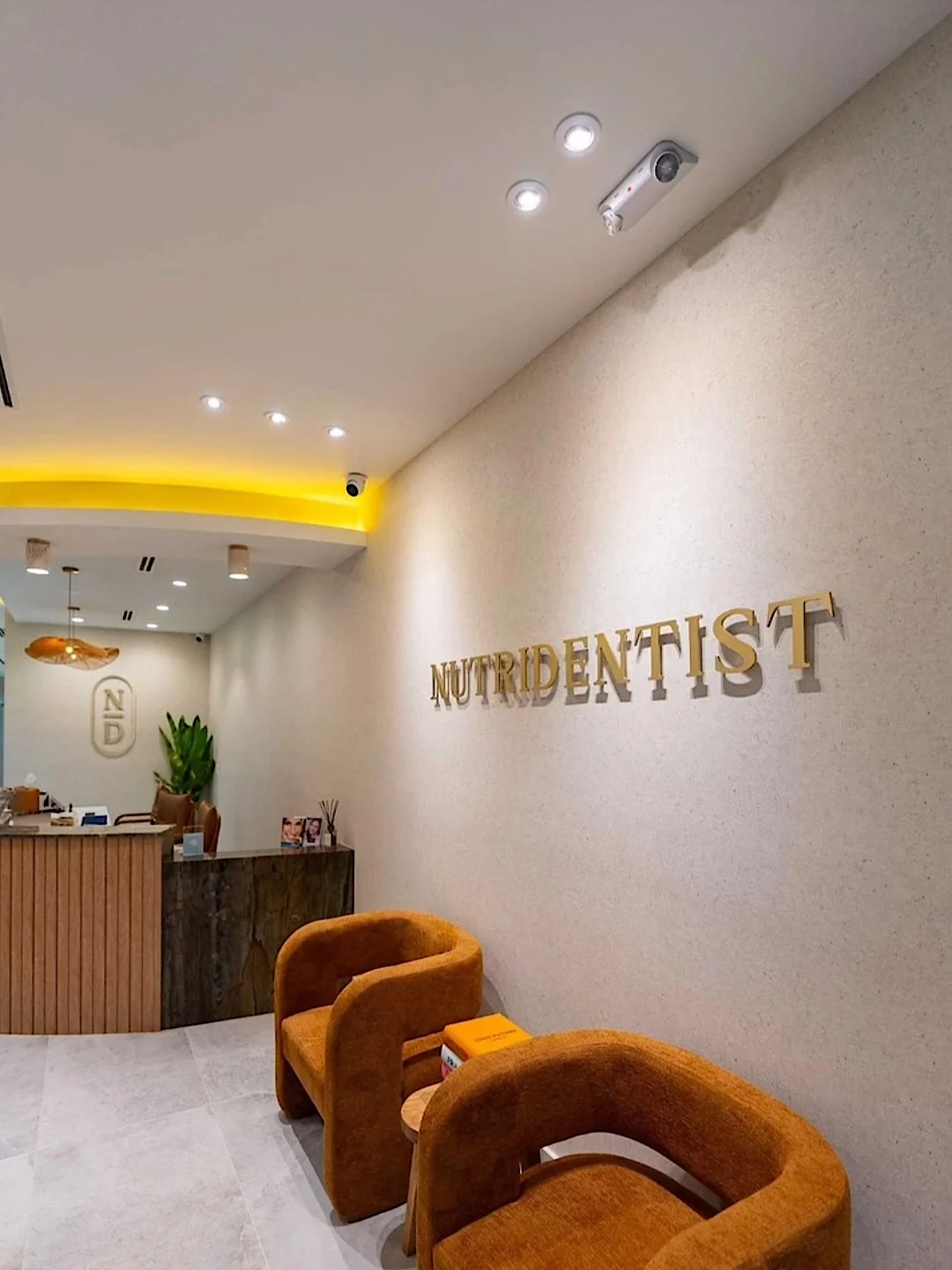 Nutridentist — Lobby con letras doradas NUTRIDENTIST sobre una pared texturizada, butacas color óxido y un mostrador de listones de madera