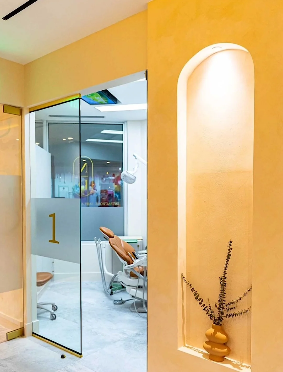 Nutridentist — Puerta de vidrio con el número 1 que da a una sala de tratamiento, junto a un nicho en arco iluminado con una rama seca