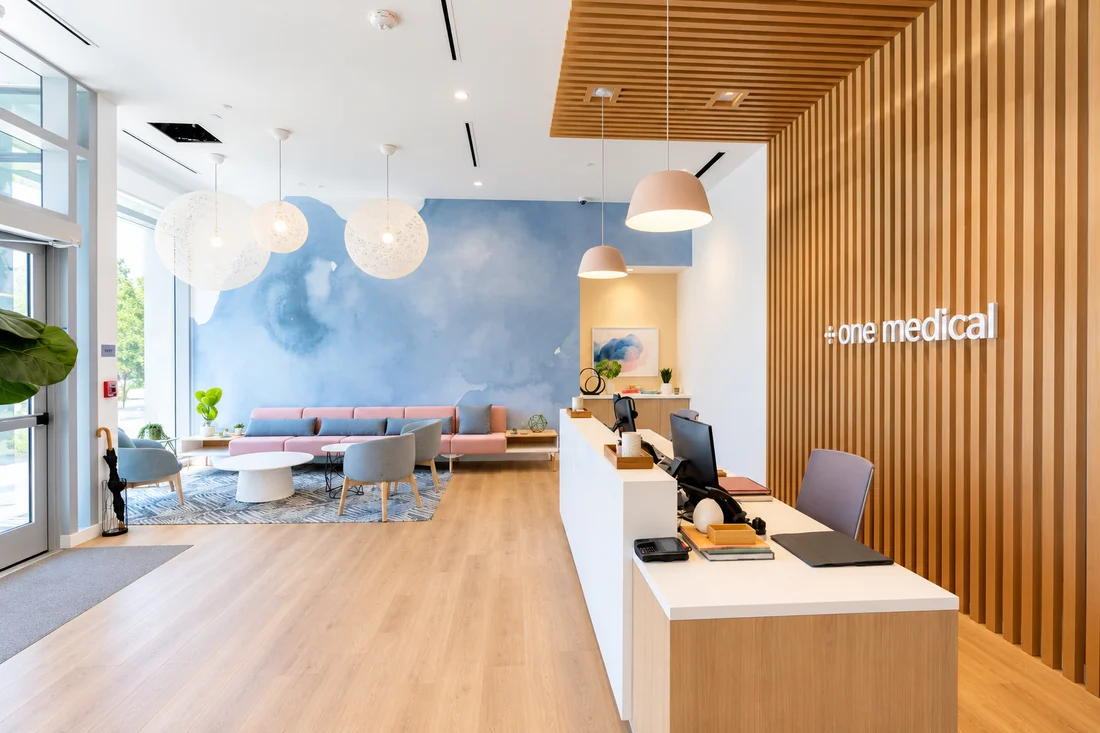 One Medical — Reception desk beside a timber-slat wall with a lettered sign, facing pink seating under a blue watercolour wall