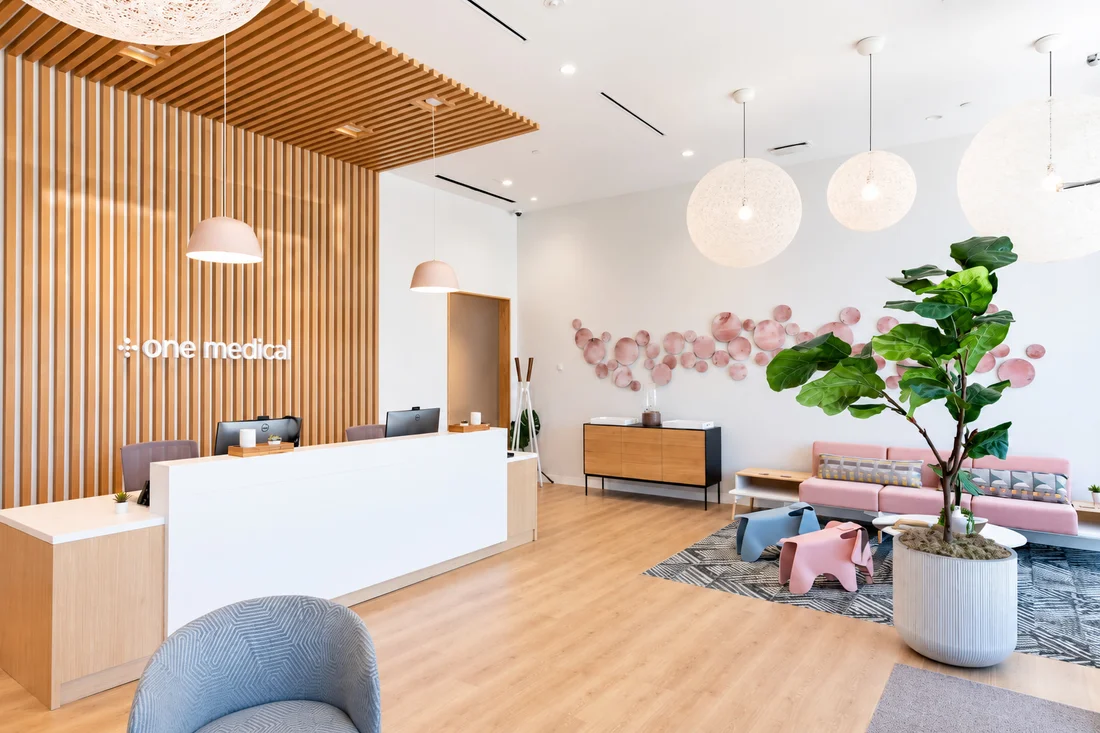 One Medical — Reception desk in front of a timber-slat wall, with pink discs across a white wall and a fiddle-leaf fig by the seating