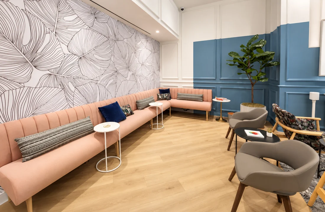 One Medical — Long peach banquette under a wall of oversized line-drawn leaves, with blue panelling and grey armchairs