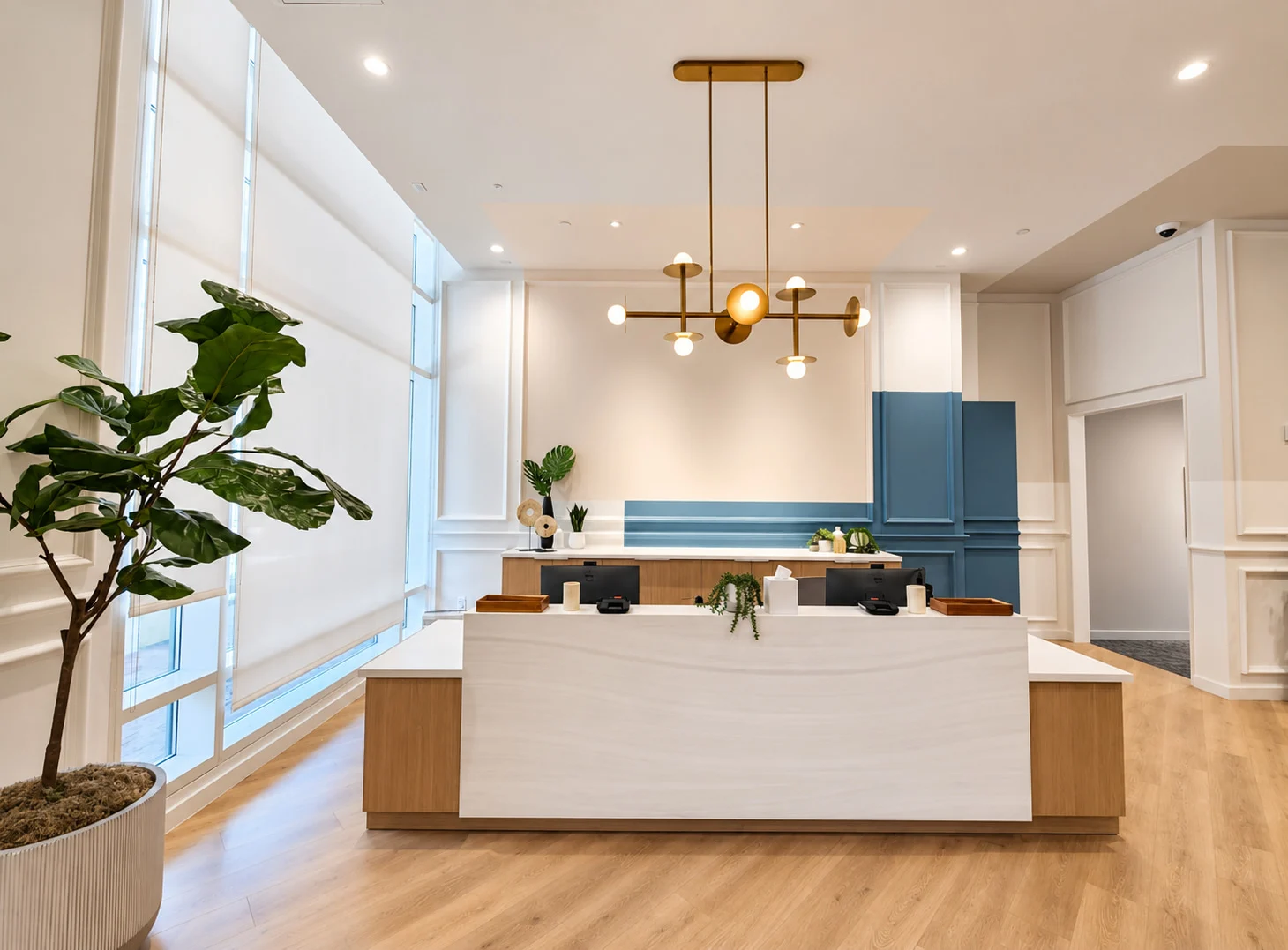 One Medical — Pale reception desk with timber sides under a brass chandelier, with white wall panelling and a block of blue