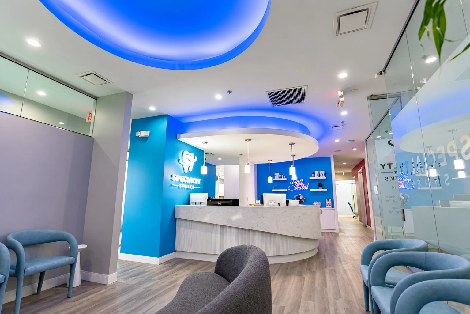 Specialty Smiles Orthodontics — Reception with a curved white desk, bright blue walls and a blue-lit ceiling cove above light blue chairs