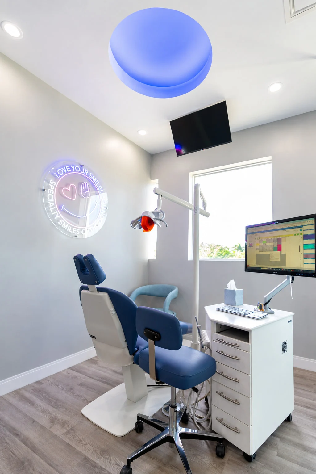 Specialty Smiles Orthodontics — Treatment room with a blue dental chair by a window and a round neon “Love your smile!” sign on the wall