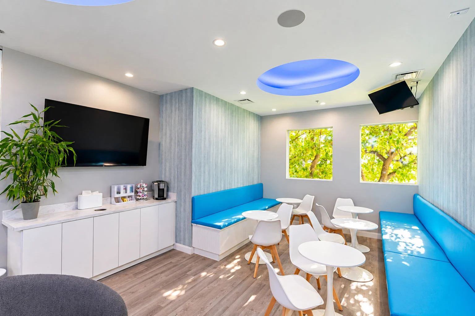 Specialty Smiles Orthodontics — Waiting lounge with bright blue banquettes, white bistro tables, a coffee counter and a blue-lit oval ceiling cove